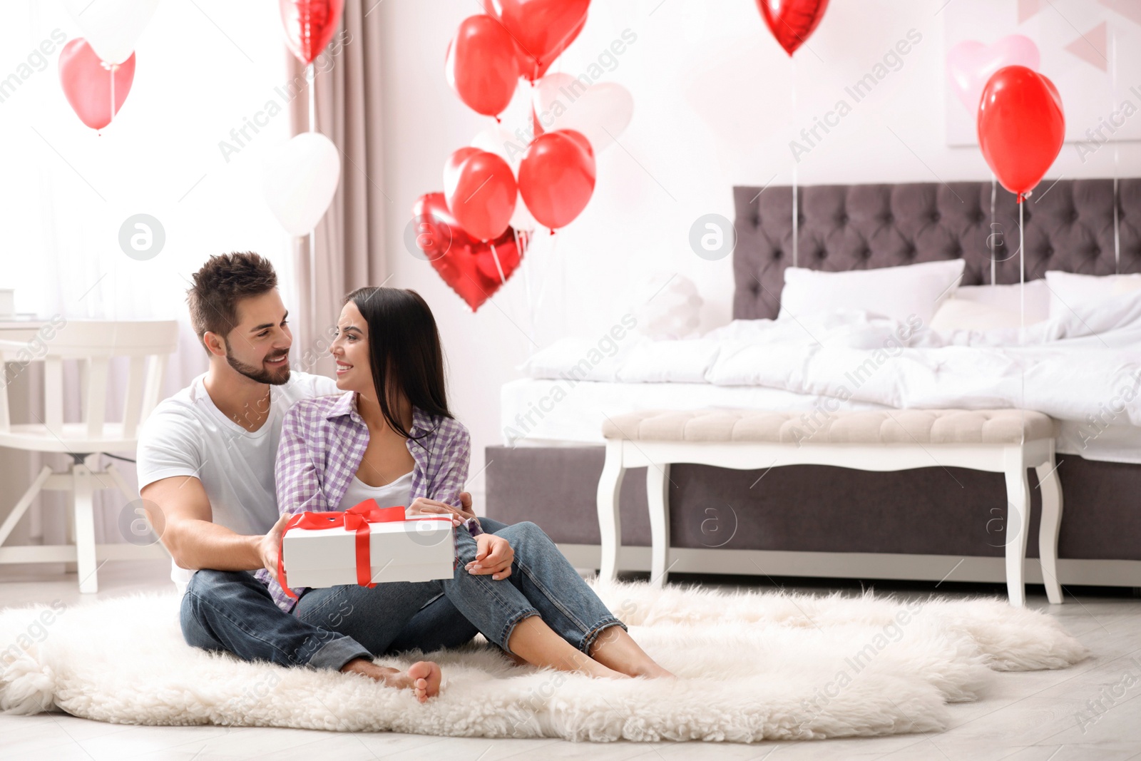 Photo of Young man presenting gift to his girlfriend in bedroom decorated with heart shaped balloons. Valentine's day celebration