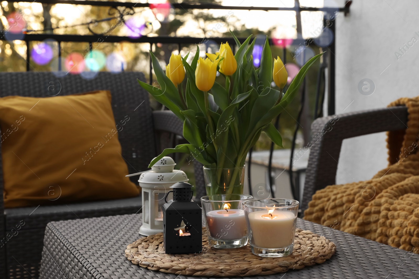 Photo of Soft pillow, blanket, burning candles and yellow tulips on rattan garden furniture outdoors