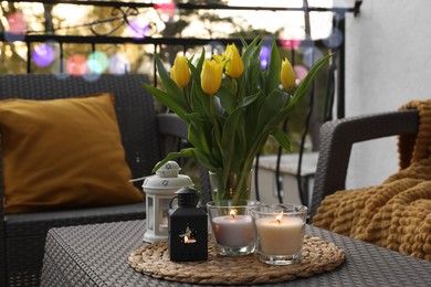 Soft pillow, blanket, burning candles and yellow tulips on rattan garden furniture outdoors