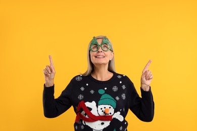 Photo of Happy senior woman in Christmas sweater and funny glasses pointing at something on orange background