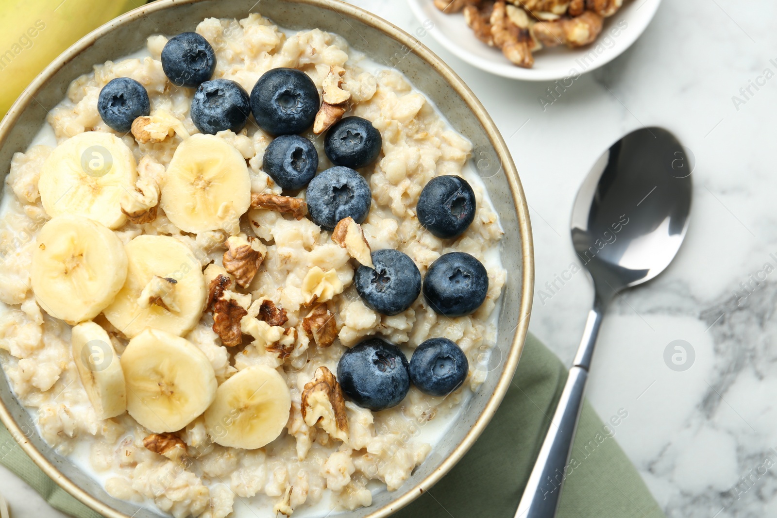 Photo of Tasty oatmeal with banana, blueberries, walnuts and milk served in bowl on white marble table, flat lay