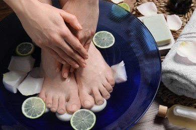 Woman soaking her feet in bowl with water, petals and lime slices on floor, closeup. Spa treatment