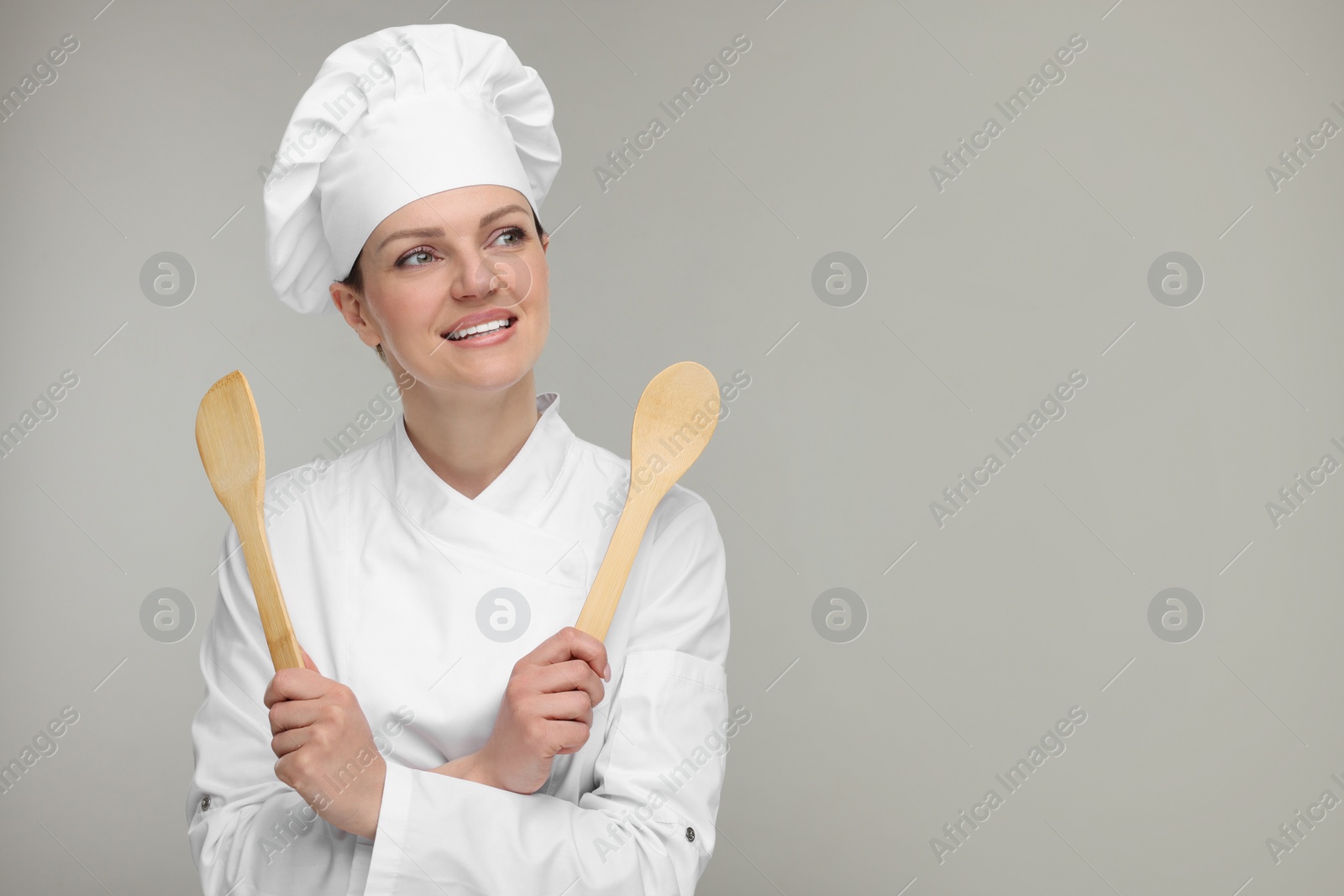 Photo of Happy woman chef in uniform holding wooden spoon and spatula on grey background, space for text
