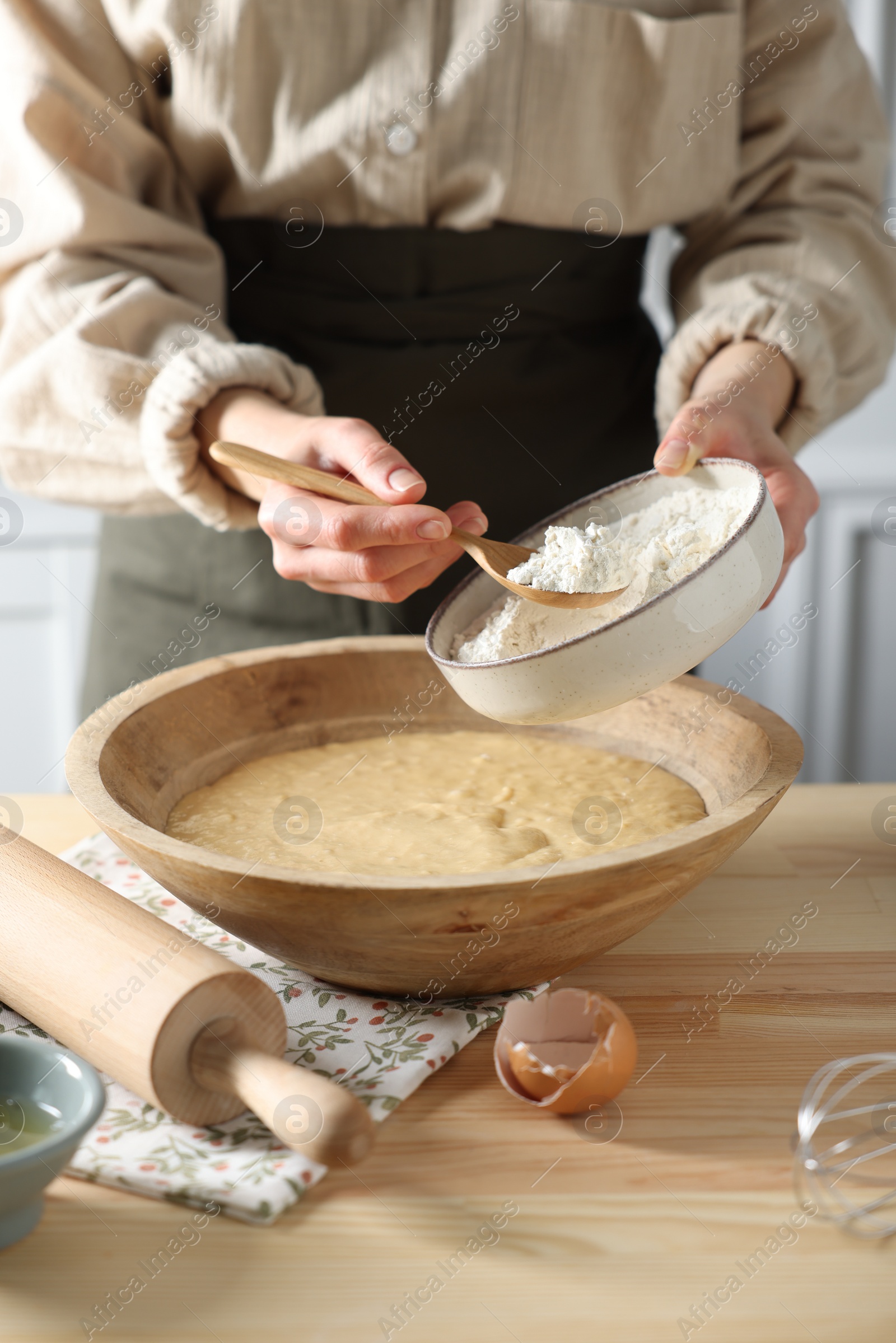 Photo of Making dough. Woman adding flour into bowl at wooden table in kitchen, closeup
