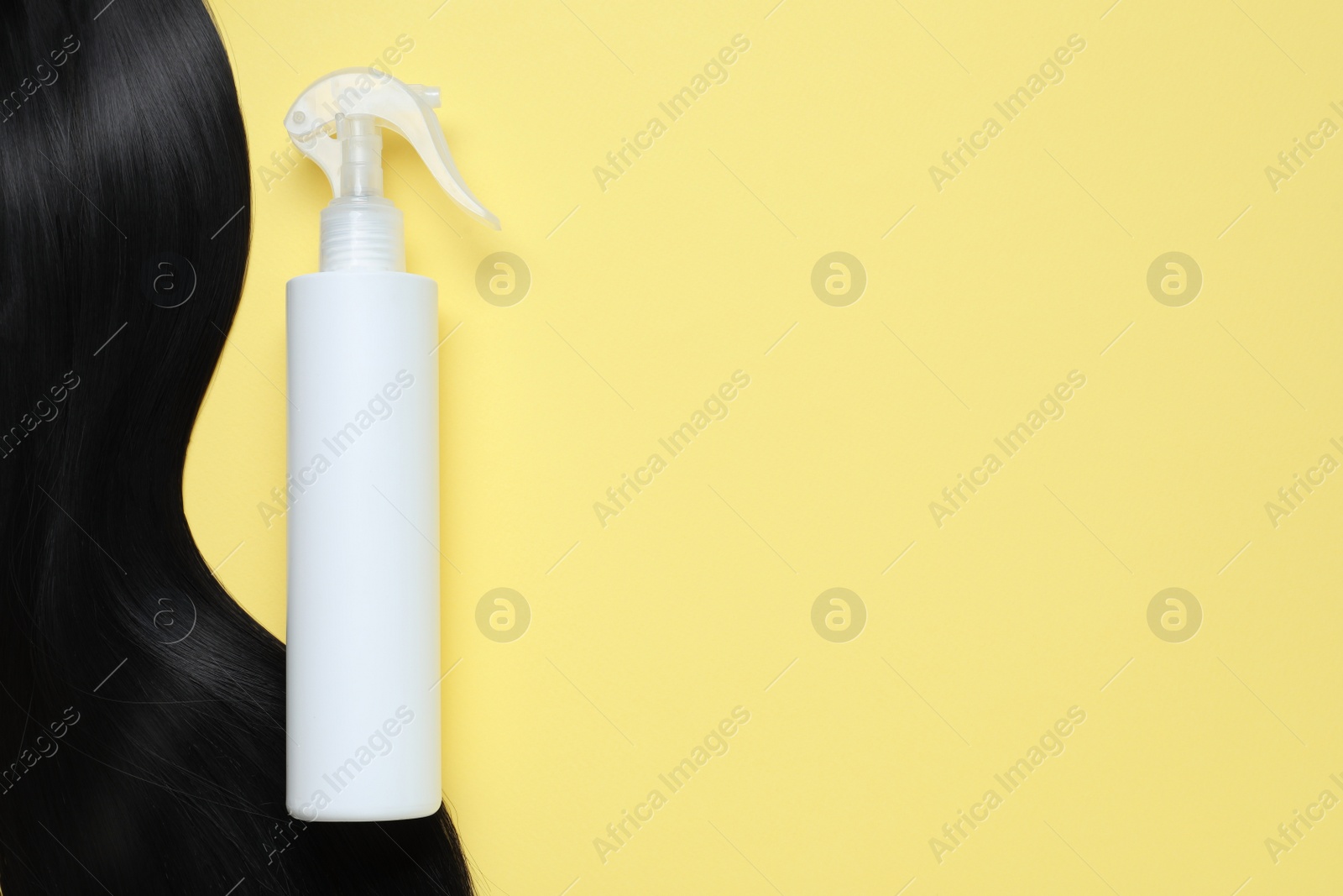 Photo of Spray bottle with thermal protection and lock of black hair on yellow background, flat lay. Space for text