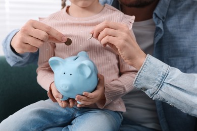 Photo of Family budget. Little girl and her parents putting coins into piggy bank at home, closeup