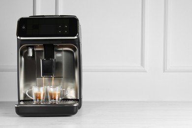 Photo of Modern espresso machine pouring coffee into glass cups on white wooden table near light wall. Space for text
