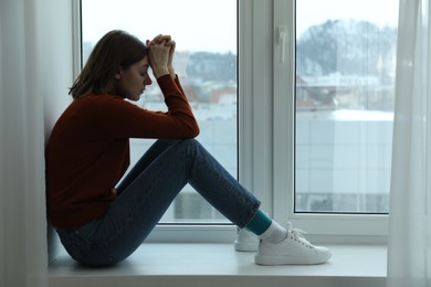 Sad young woman sitting on windowsill near window at home, space for text