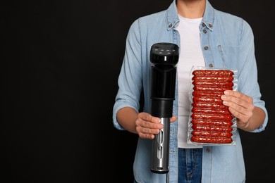 Woman holding sous vide cooker and sausages in vacuum pack on black background, closeup. Space for text