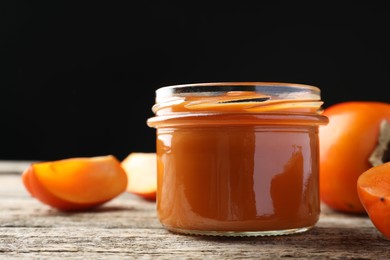 Photo of Delicious persimmon jam in glass jar and fresh fruits on wooden table against black background, closeup. Space for text