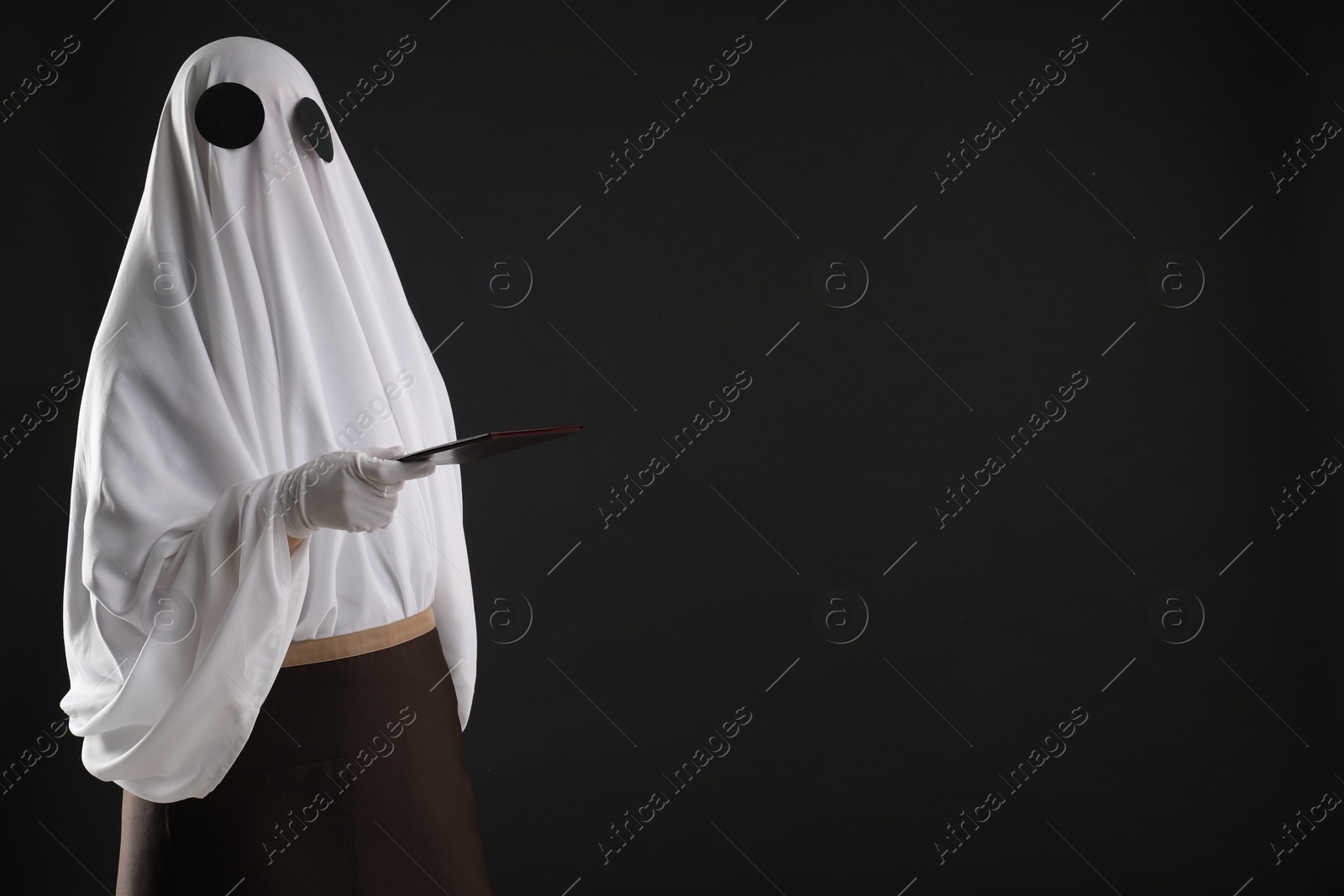 Photo of Creepy ghost. Waiter in white sheet and apron with receipt on black background, space for text