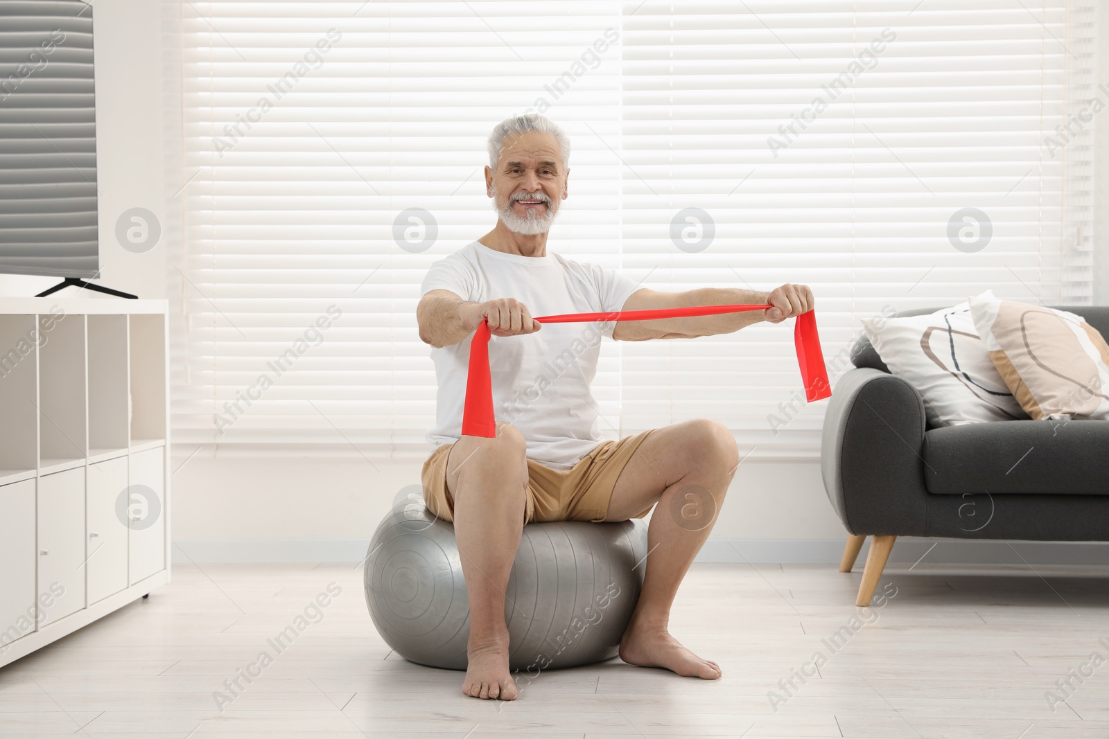 Photo of Senior man doing exercise with elastic resistance band on fitness ball at home