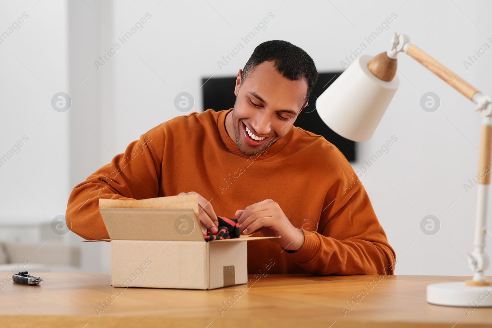 Photo of Happy young man opening parcel at table indoors. Internet shopping