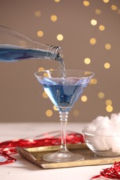 Cocktail making. Pouring tasty alcoholic drink in glass at white wooden table against beige background with blurred lights, closeup