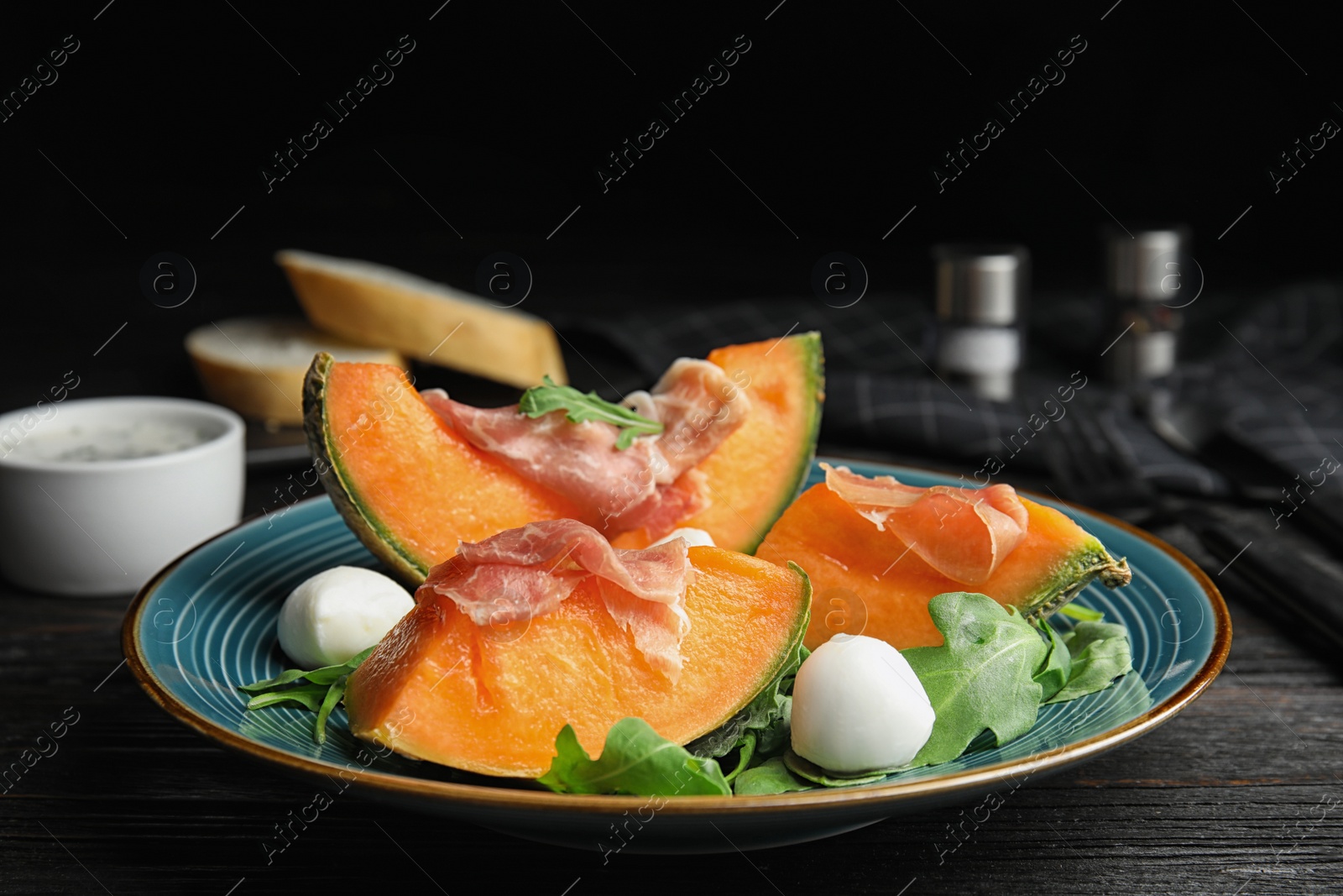 Photo of Fresh melon with prosciutto, mozzarella and arugula on black wooden table