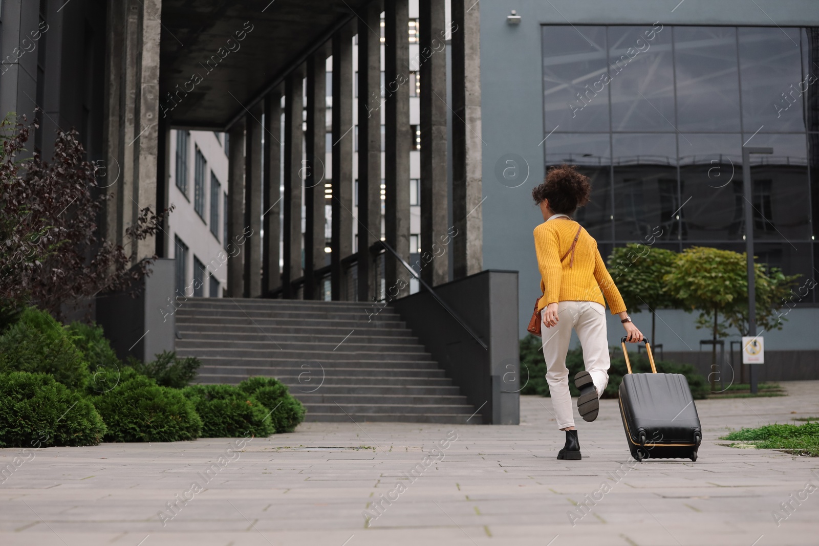 Photo of Being late. Woman with suitcase running towards building outdoors, back view