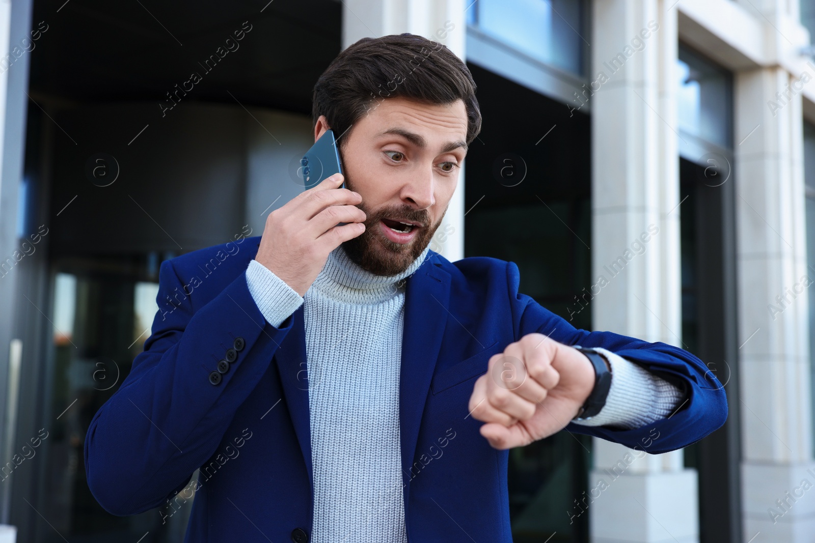 Photo of Emotional businessman talking on smartphone and looking at watch near building. Being late concept