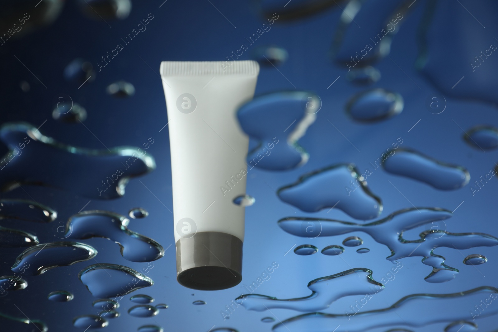 Photo of Moisturizing cream in tube on glass with water drops against blue background, low angle view. Space for text