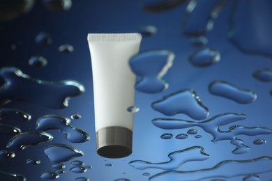 Photo of Moisturizing cream in tube on glass with water drops against blue background, low angle view. Space for text