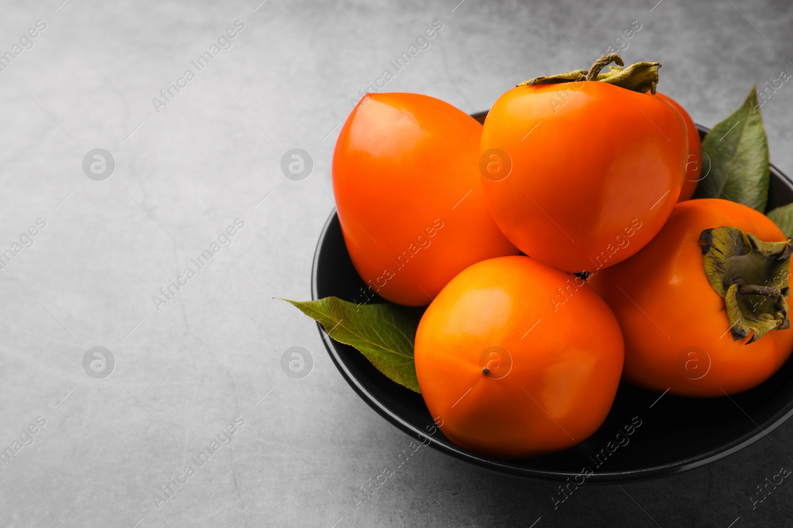 Photo of Delicious ripe persimmons in bowl on light gray textured table, space for text