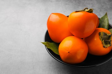 Delicious ripe persimmons in bowl on light gray textured table, space for text