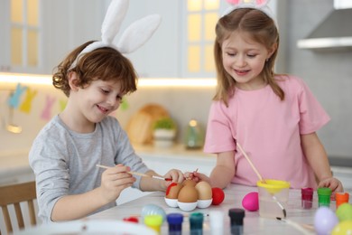 Photo of Easter celebration. Cute children with bunny ears painting eggs at white marble table in kitchen