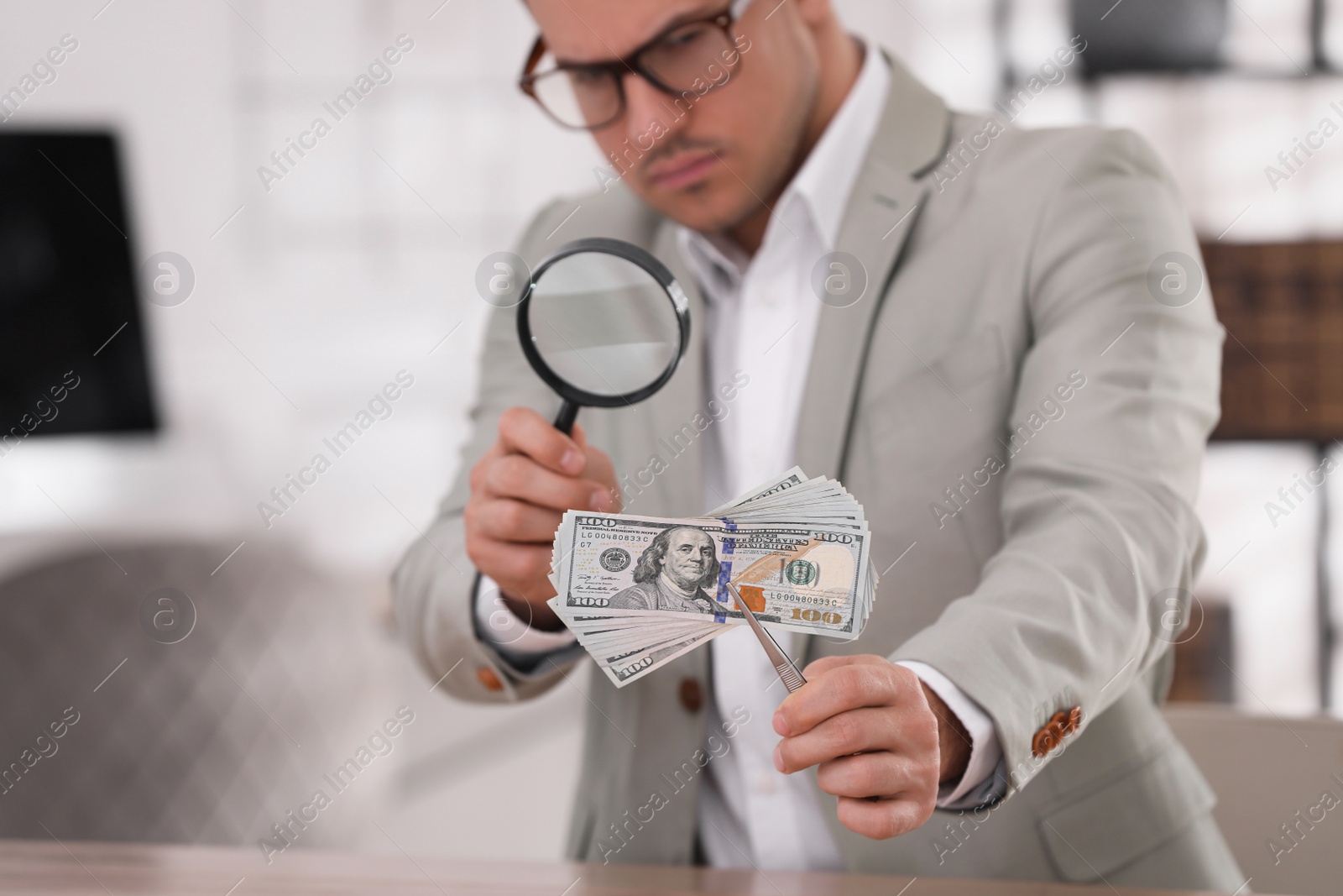 Photo of Expert authenticating 100 dollar banknotes with magnifying glass at table in office, focus on hand. Fake money concept