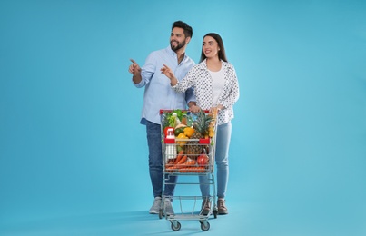 Young couple with shopping cart full of groceries on light blue background