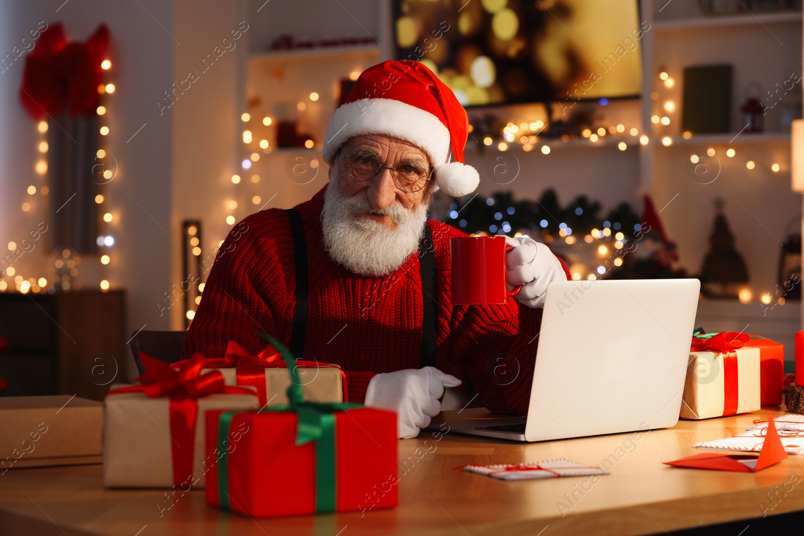 Photo of Santa Claus using laptop and drinking hot beverage at his workplace in room decorated for Christmas