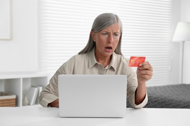 Photo of Shocked woman looking at credit card near laptop at table indoors. Be careful - fraud