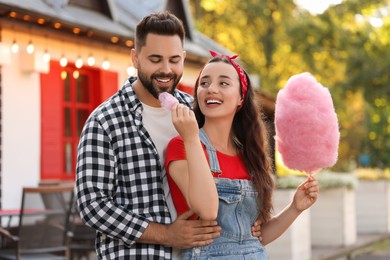 Photo of Happy young woman treating her boyfriend with cotton candy outdoors
