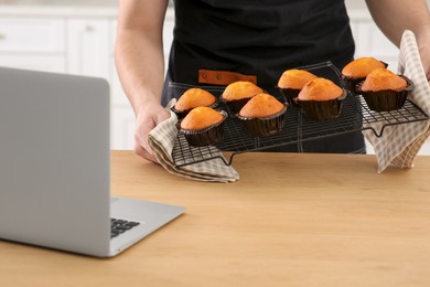 Man holding muffins near laptop at wooden table indoors, closeup. Time for hobby
