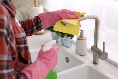 Woman with spray bottle and microfiber cloth cleaning water tap in kitchen, closeup