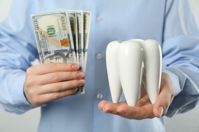 Woman holding ceramic model of tooth and dollar banknotes on light background, closeup. Expensive treatment