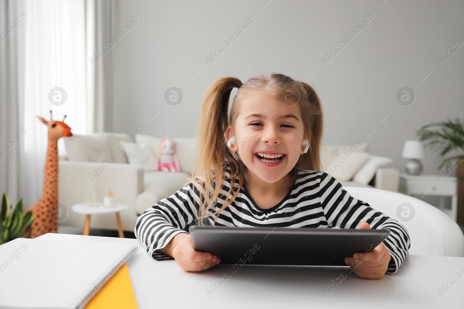 Photo of Adorable little girl doing homework with tablet and earphones at table indoors