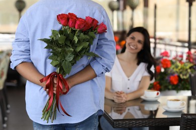 International dating. Man hiding bouquet of roses for his beloved woman in restaurant, selective focus