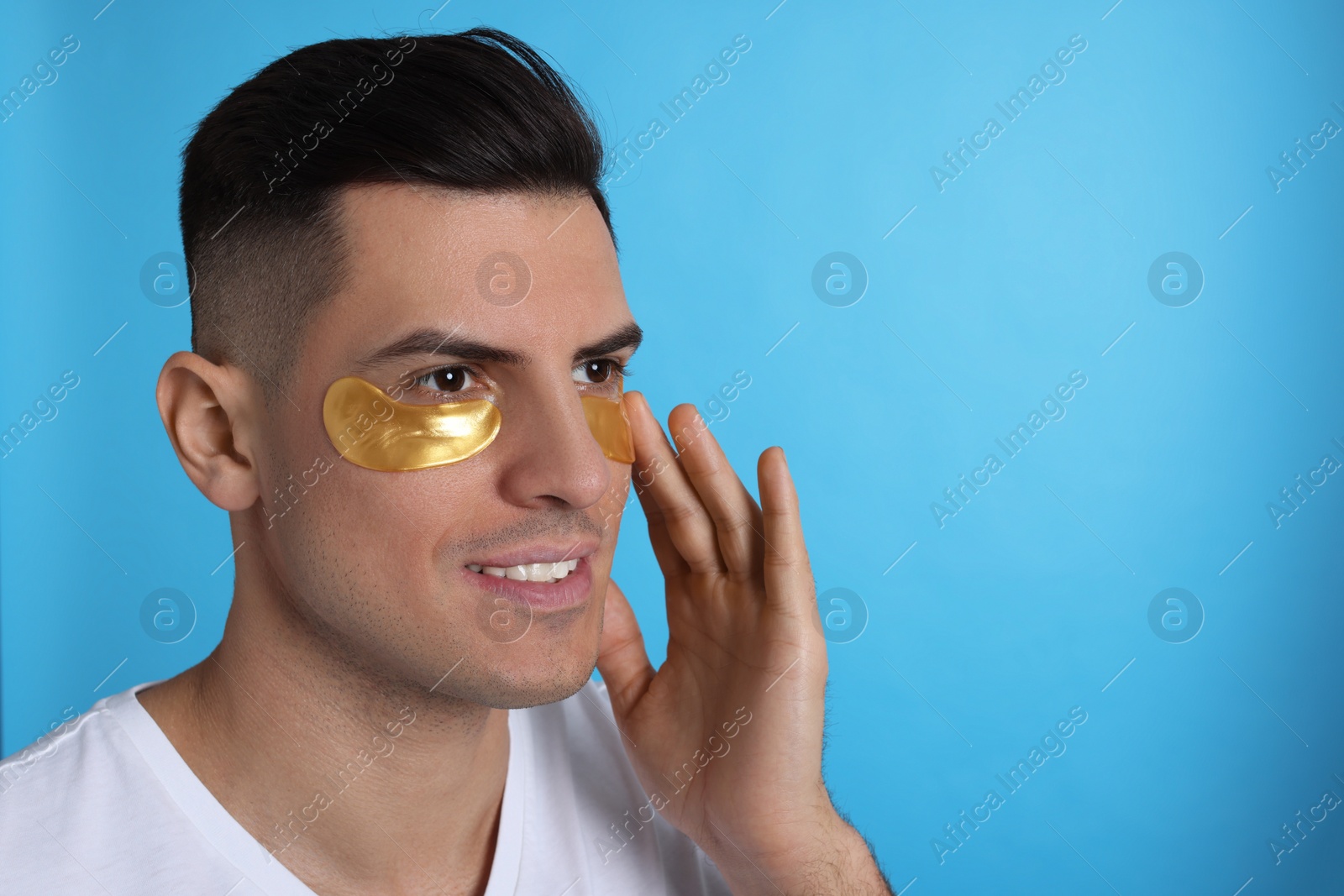 Photo of Man applying golden under eye patch on light blue background. Space for text
