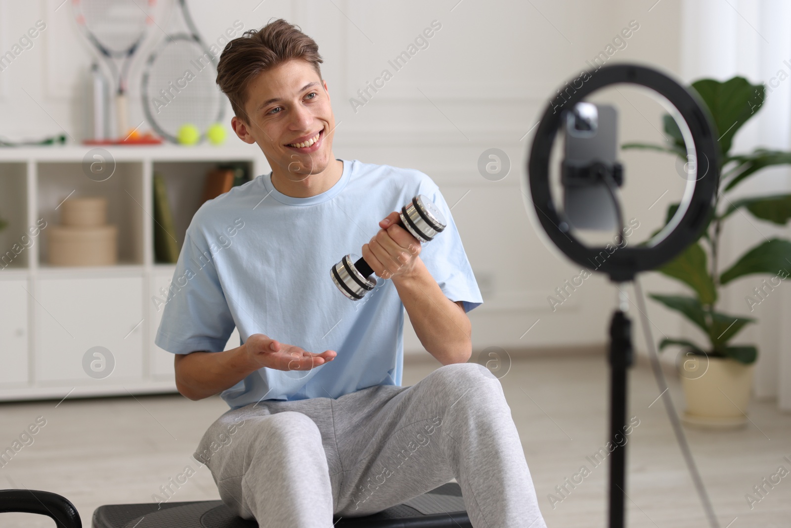 Photo of Smiling sports blogger holding dumbbell while streaming online fitness lesson with smartphone at home