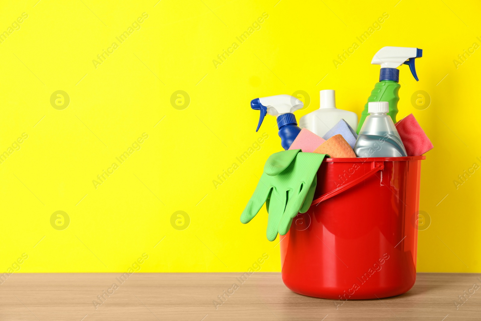 Photo of Bucket with different cleaning supplies on wooden floor near yellow wall. Space for text