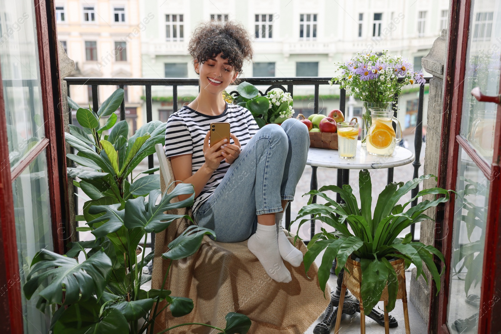 Photo of Beautiful young woman using smartphone at table on balcony with houseplants