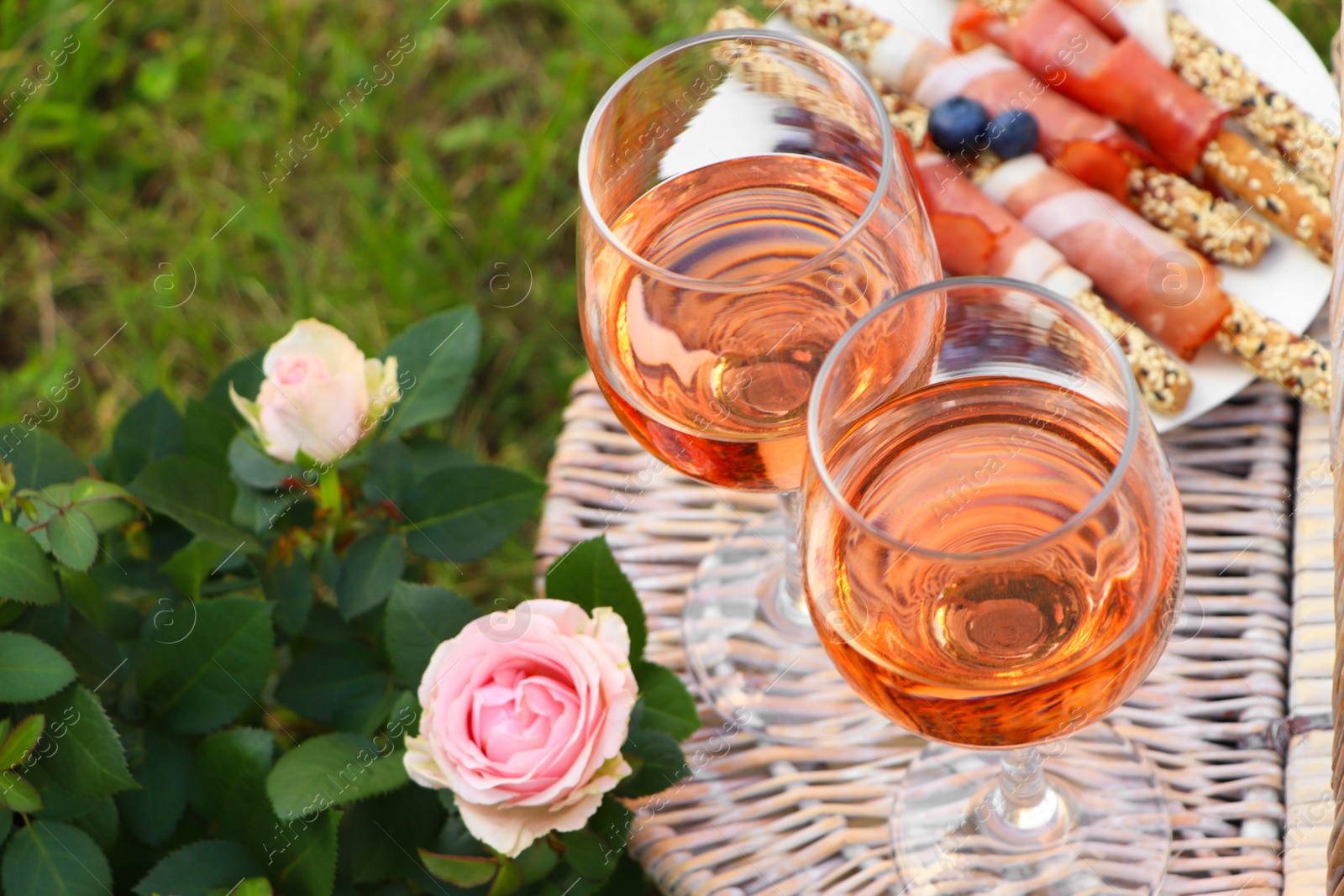 Photo of Flowers near glasses of delicious rose wine and food on picnic basket outdoors, closeup