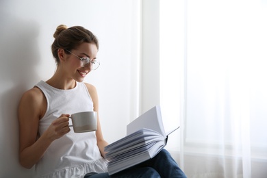 Young woman with cup of coffee reading book at home, space for text