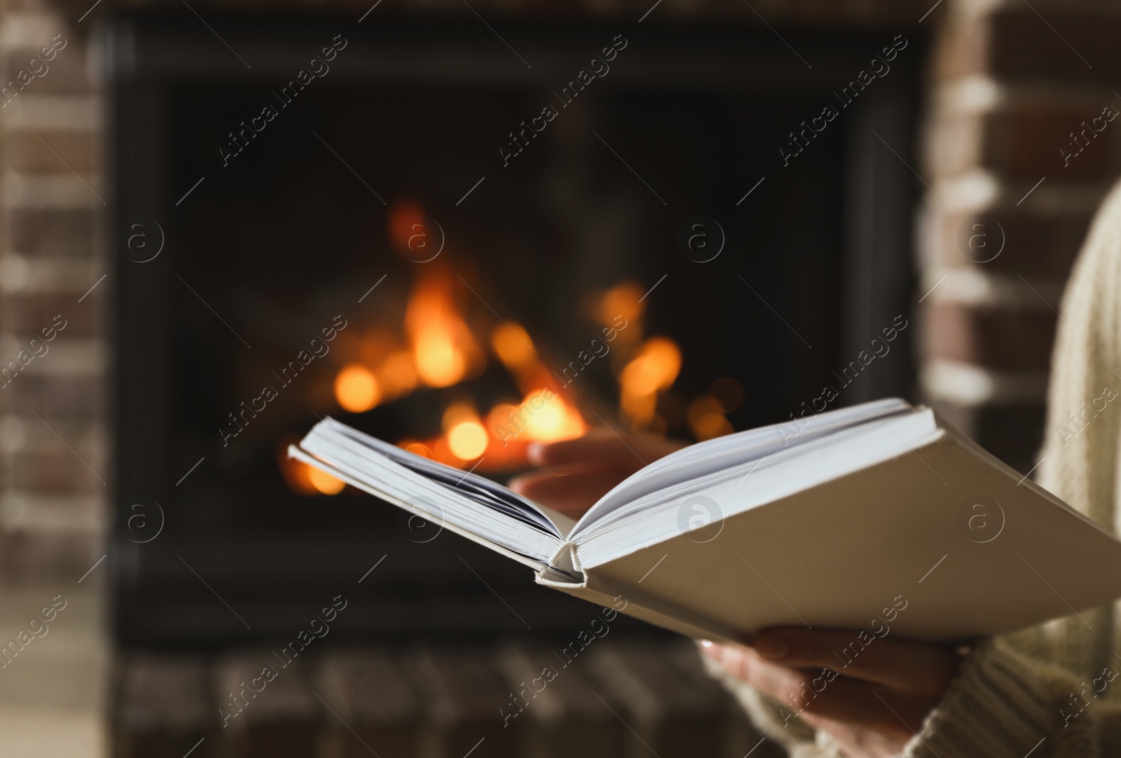 Photo of Woman reading book near burning fireplace at home, closeup