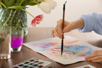 Photo of Woman painting flowers with watercolor at wooden table indoors, closeup. Creative artwork
