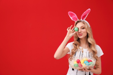Photo of Beautiful young woman in bunny ears headband with Easter eggs on color background, space for text