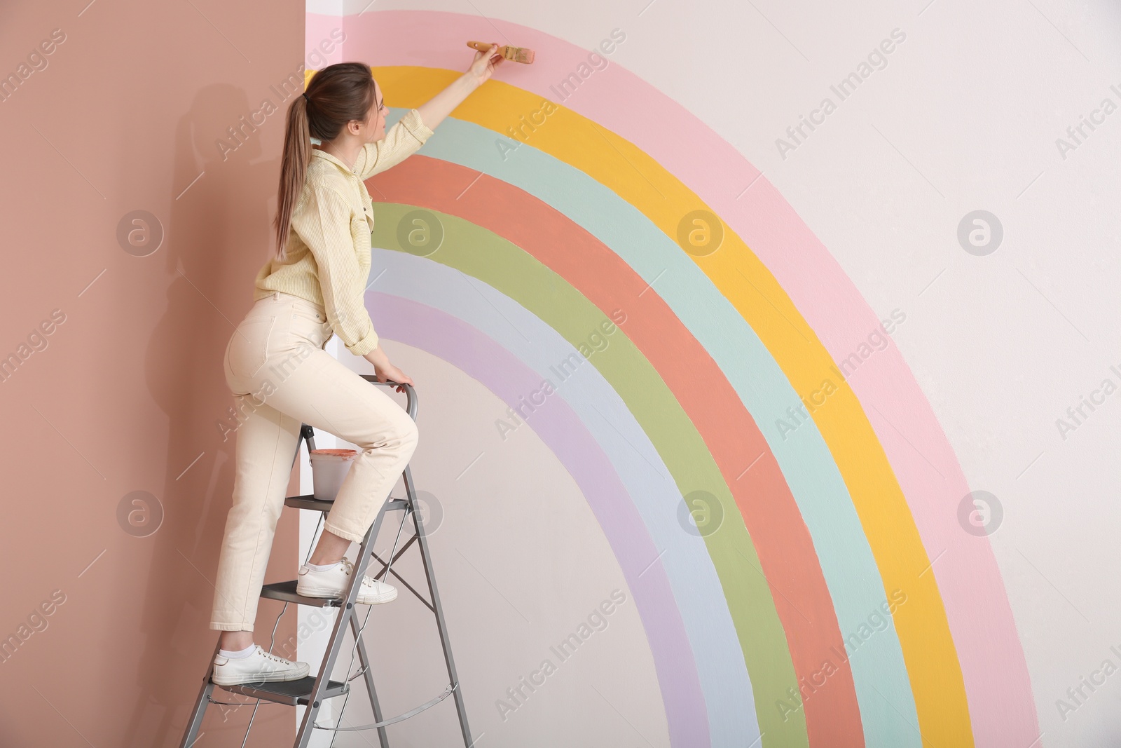 Photo of Young woman painting rainbow on white wall indoors, space for text