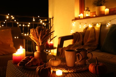 Photo of Rattan furniture, cups, fairy lights, burning candles and other autumn decor on outdoor terrace at night
