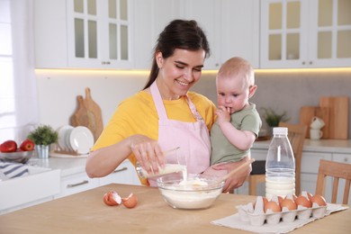 Photo of Happy young woman holding her cute little baby while making dough in kitchen