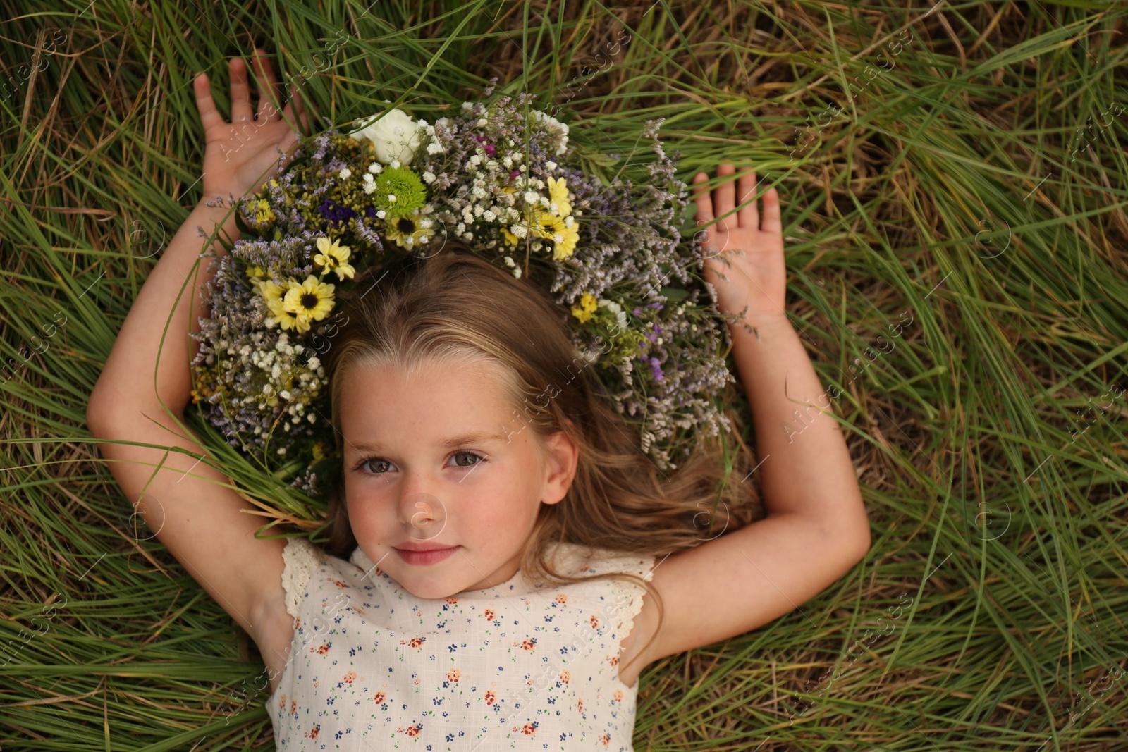 Photo of Cute little girl wearing wreath made of beautiful flowers on green grass, top view