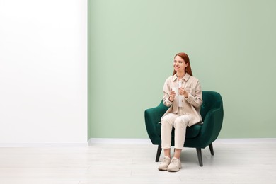 Happy young woman with cup of drink sitting in armchair indoors. Space for text